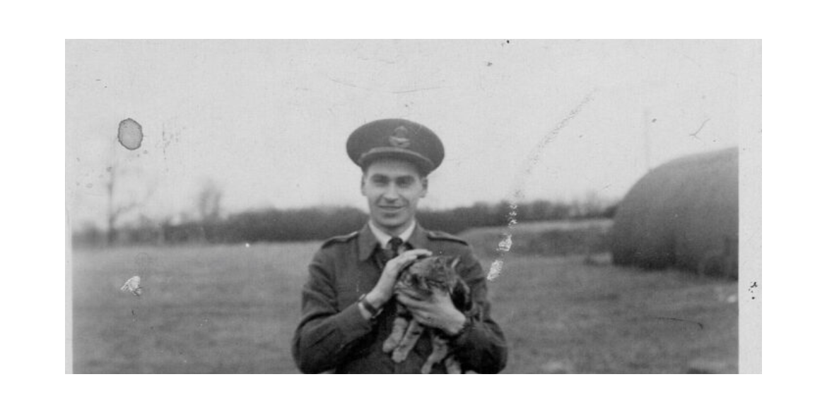 Smiling man in a military uniform holding a small cat outdoors in a field.