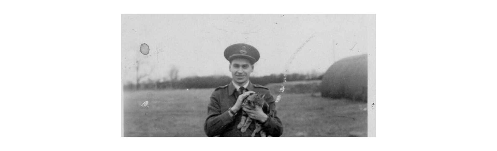 Smiling man in a military uniform holding a small cat outdoors in a field.
