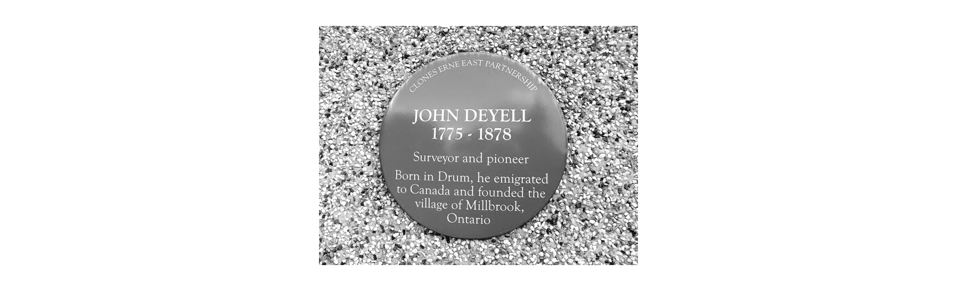 Circular commemorative plaque on gravel: 'John Deyell 1775–1878' with text about a surveyor and pioneer and the village of Millbrook, Ontario.