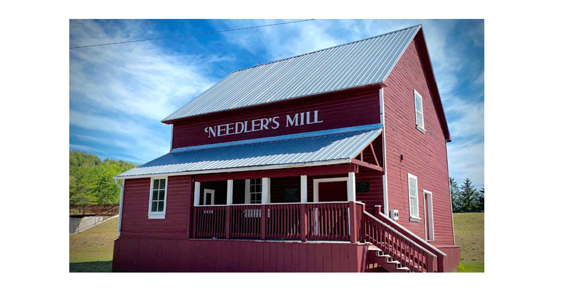 Red two-story mill building labeled 'Needler's Mill' with a white-trimmed porch and metal roof on a sunny day, blue sky above.
