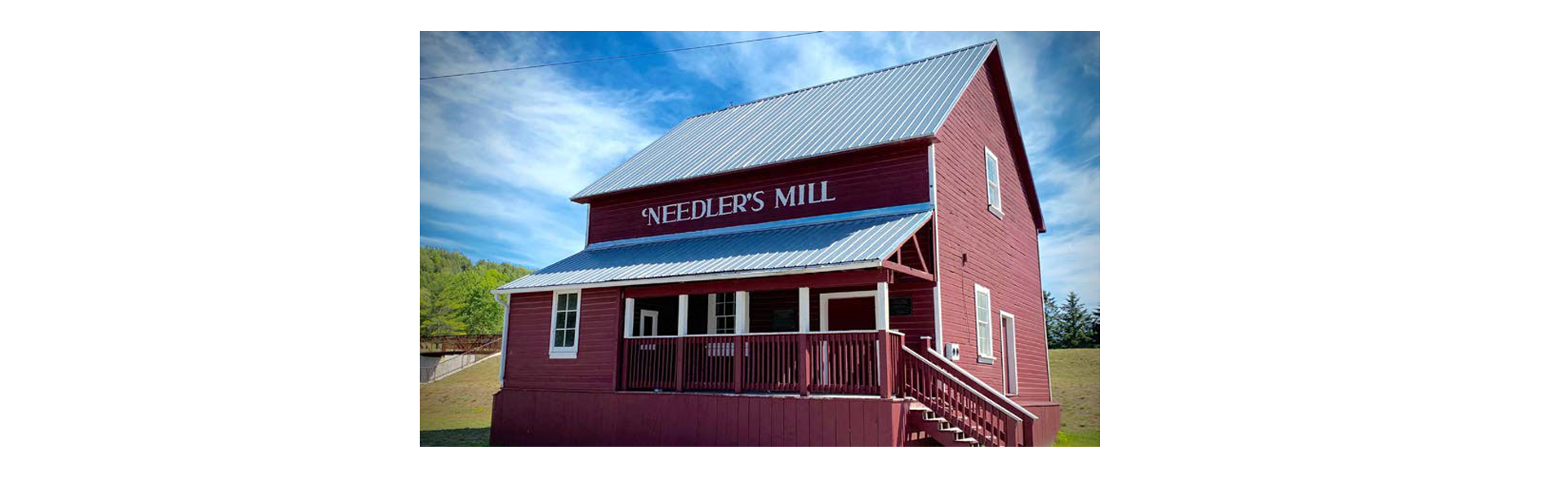 Red two-story mill building labeled 'Needler's Mill' with a white-trimmed porch and metal roof on a sunny day, blue sky above.