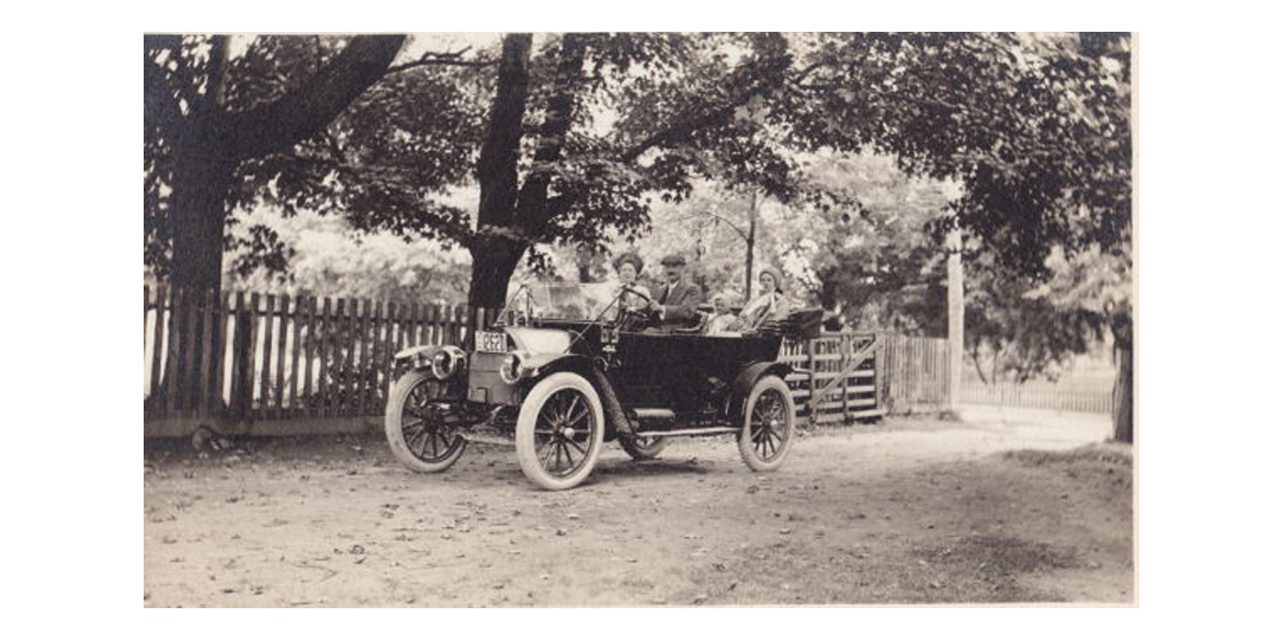 Vintage open-top car with four passengers on a dirt road beside a wooden fence under trees in a black-and-white photo.
