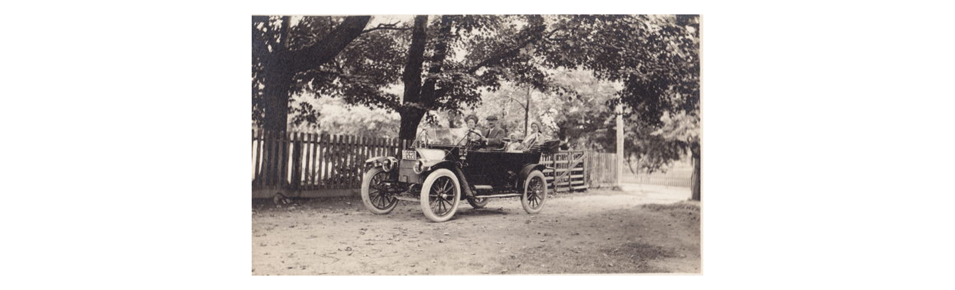 Vintage open-top car with four passengers on a dirt road beside a wooden fence under trees in a black-and-white photo.