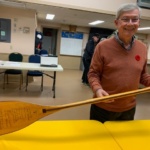 Older man in an orange sweater smiles while holding a long wooden oar with writing on it, indoors at a community center.
