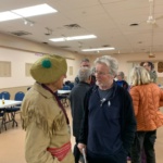 Older man in a dark blue zip-up sweater chats with a woman wearing a tan fringed jacket and a green hat in a community hall with tables in the background.