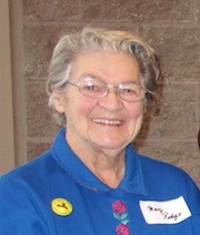 Smiling elderly woman with gray hair and glasses, wearing a blue jacket and badge at a public event.
