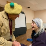 Elderly man in a fringed tan jacket and yellow knit hat with a green pom-pom shakes hands with an elderly woman in a purple sweater and patterned headscarf, both smiling indoors.