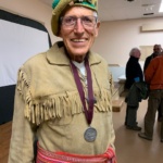 Older man wearing a fringe suede jacket, decorated hat, and a silver medal on a purple lanyard at an indoor event.
