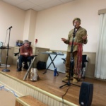Man in rustic traditional attire speaks into a microphone on a small stage as another man sits nearby; a log and axe lie on the floor beside them.