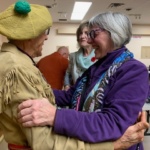 Two elderly women share a warm hug and laugh in a community room, with others chatting in the background.