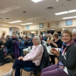 Audience of mostly seniors seated in a community hall, clapping during an event or performance.