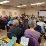Crowded community hall with seniors listening to a speaker at a podium and a projection screen at the front.