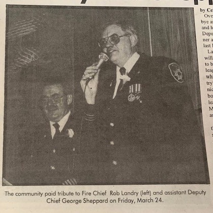 Black-and-white photo of Fire Chief Rob Landry (left) speaking into a microphone at a community tribute with Assistant Deputy Chief George Sheppard nearby.