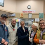 Five smiling seniors stand together in a community room near a kitchen area, wearing red pins; a 'No Drinks On Dance Floor' sign is visible in the background.