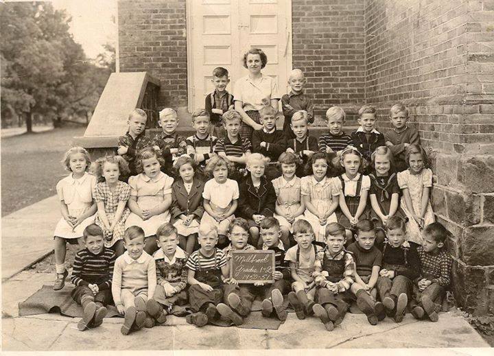 Group of schoolchildren and a teacher posing on stone steps outside a brick building; front row holds a chalkboard for the class photo period.